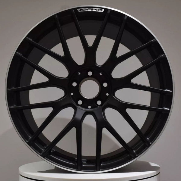 A black, multi-spoke AMG car rim is displayed upright against a neutral background, resting on a white surface.