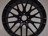 A black alloy wheel rests upright on a white surface. The wheel features a multi-spoke design and a label at the top reading 
