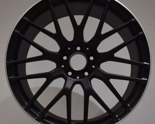 A black alloy wheel rests upright on a white surface. The wheel features a multi-spoke design and a label at the top reading "AMG." The background is plain gray.