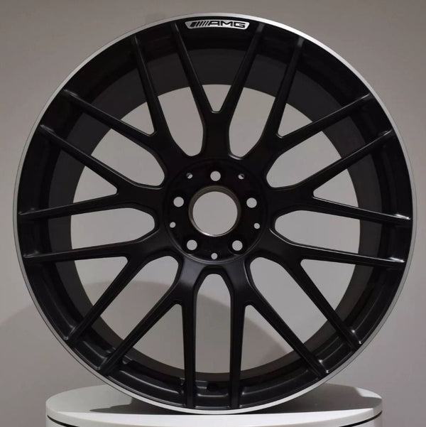 A black alloy wheel rests upright on a white surface. The wheel features a multi-spoke design and a label at the top reading "AMG." The background is plain gray.