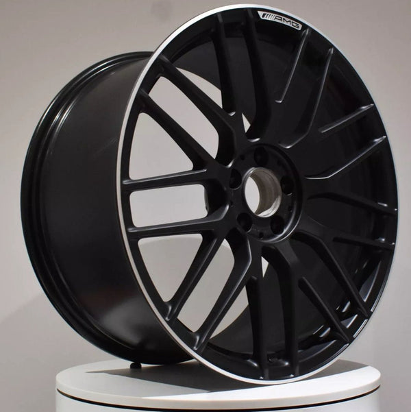 A black alloy wheel with a multi-spoke design resting on a white surface. It features an "AMG" logo on the rim within an indoor setting.