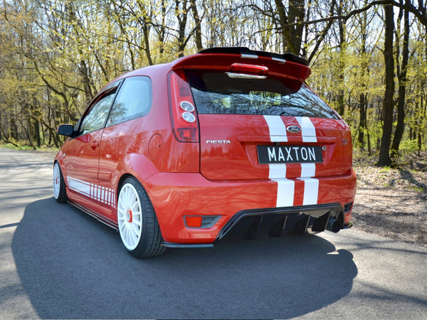 A red Ford Fiesta with white stripes is stationary on a tree-lined road. The license plate reads "MAXTON."