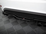 Car's rear diffuser with dual exhaust pipes, showcasing a black honeycomb pattern and logo, resting on a textured gray mat in a garage setting.