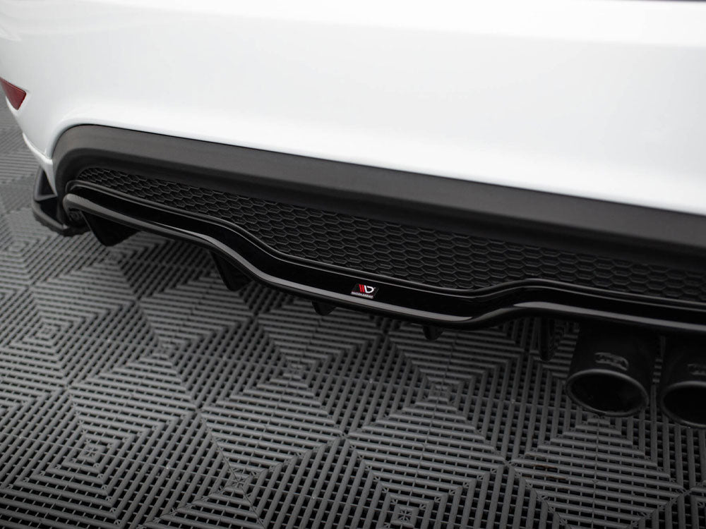 Car's rear diffuser with dual exhaust pipes, showcasing a black honeycomb pattern and logo, resting on a textured gray mat in a garage setting.