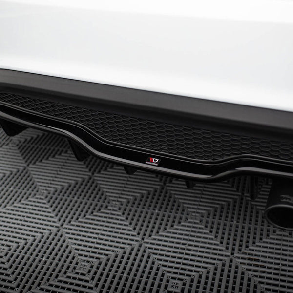 Car's rear diffuser with dual exhaust pipes, showcasing a black honeycomb pattern and logo, resting on a textured gray mat in a garage setting.