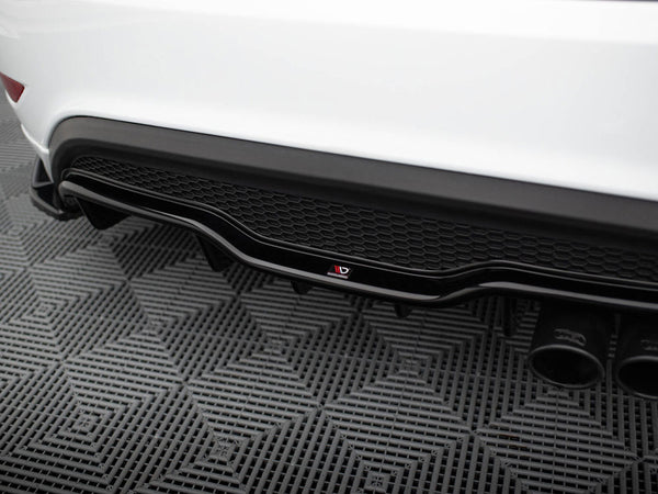 Car's rear diffuser with dual exhaust pipes, showcasing a black honeycomb pattern and logo, resting on a textured gray mat in a garage setting.