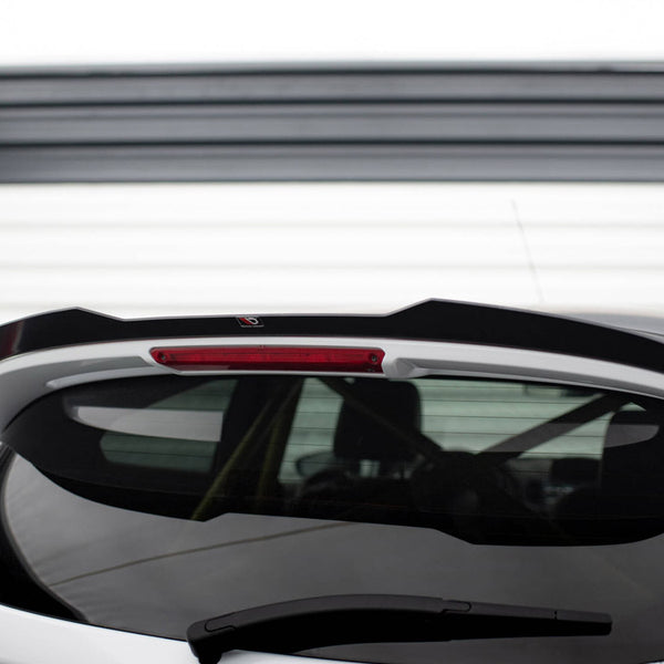 A white car's rear hatch with a black spoiler, featuring a centered red brake light, is parked in front of a white garage door with horizontal panels.