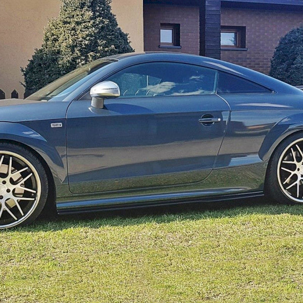 A sleek, dark gray sports car with large alloy wheels is parked on a grassy lawn in front of a house, bordered by evergreen shrubs and a wooden fence.