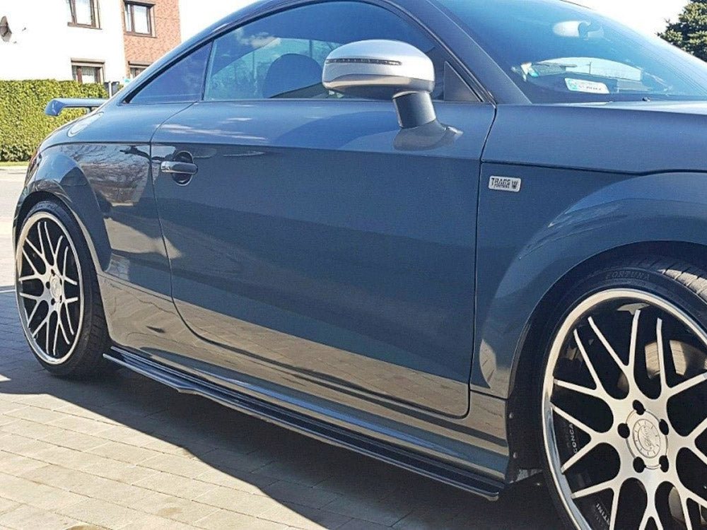 A dark blue sports car parked on a sunlit driveway, featuring large black alloy wheels with silver spokes and a "Traffic W" badge on its side.
