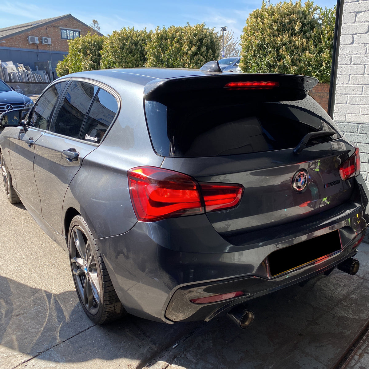 A sleek, dark gray car is parked outdoors, featuring prominent red tail lights and a BMW badge. Text on the rear reads "M135I" and "xDrive." Nearby, bushes and buildings are visible.