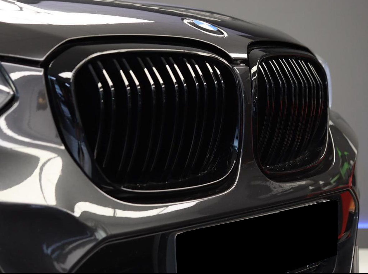 The car's front features a glossy black grille with vertical slats, displaying the BMW emblem on the hood, set against a blurred indoor background.