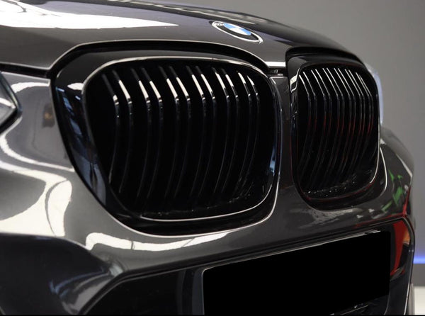 The car's front features a glossy black grille with vertical slats, displaying the BMW emblem on the hood, set against a blurred indoor background.