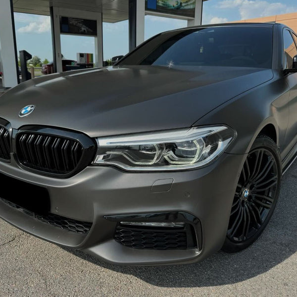 A sleek, matte gray BMW sedan is parked at a gas station with fuel pumps visible in the background under a clear sky.