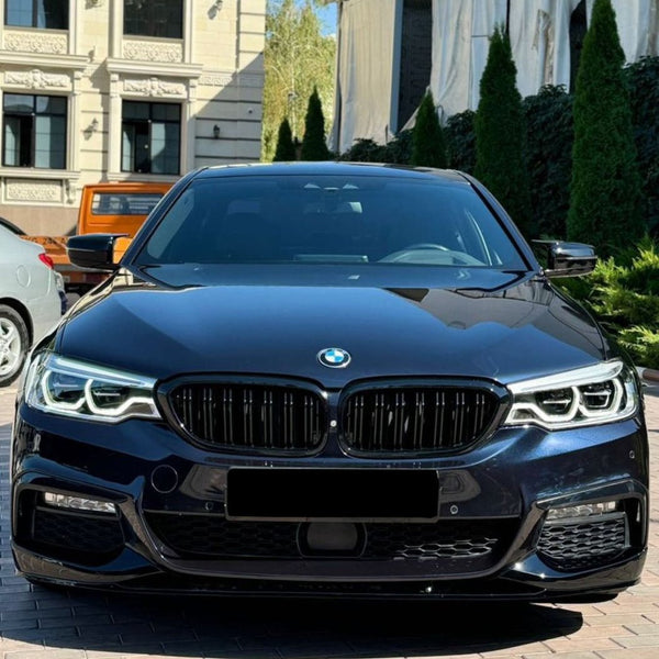 A sleek black car with prominent headlights is parked on a brick driveway. It is flanked by two other cars and bordered by tall green shrubs and buildings in the background.