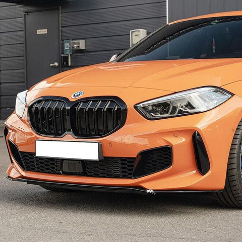 A bright orange BMW car is parked, displaying a prominent front grille and sleek headlights on a street with a building entrance nearby. The license plate is white.