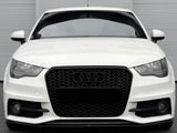 A white car with a black honeycomb grille and Audi emblem is stationary, displayed front-facing against a light gray, vertically paneled wall in a parking area.