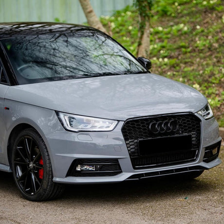 A grey car with a black grille and wheels is parked on a concrete surface, surrounded by green foliage and trees. The headlights are designed in a modern style.