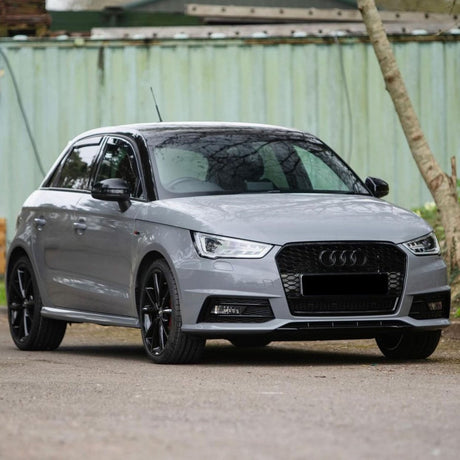 A grey hatchback car is parked on a road, featuring sleek black wheels and an emblem on the front. It is positioned in front of a green metal fence and trees.