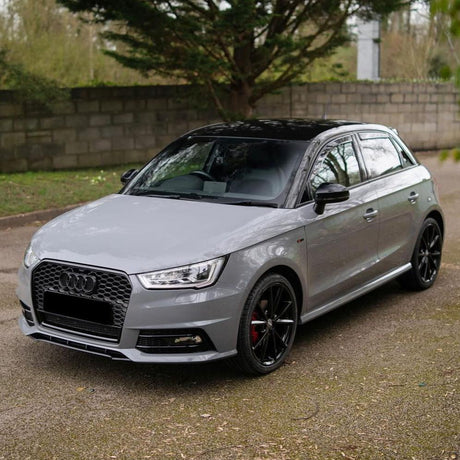 A gray car with a black roof, parked on a paved path, surrounded by greenery and a stone wall in the background.