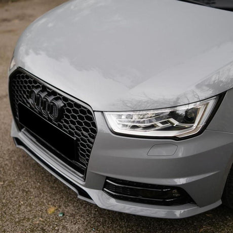 A grey car's front end with a honeycomb grille, distinctive headlights, and no visible license plate, parked on a textured pavement surface outdoors.
