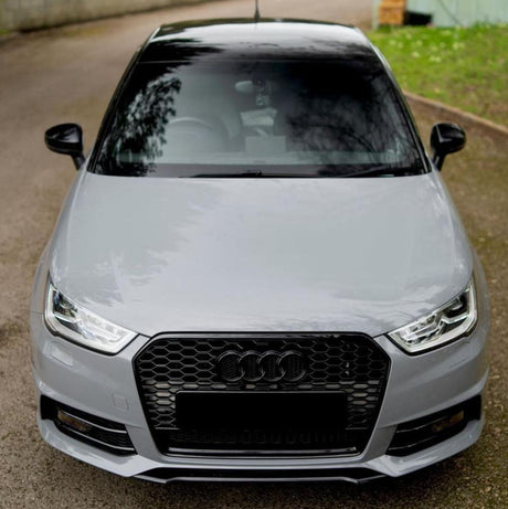 A silver car with a hexagonal grille and prominent headlights is parked on a paved driveway, surrounded by a green lawn and wooden fencing.