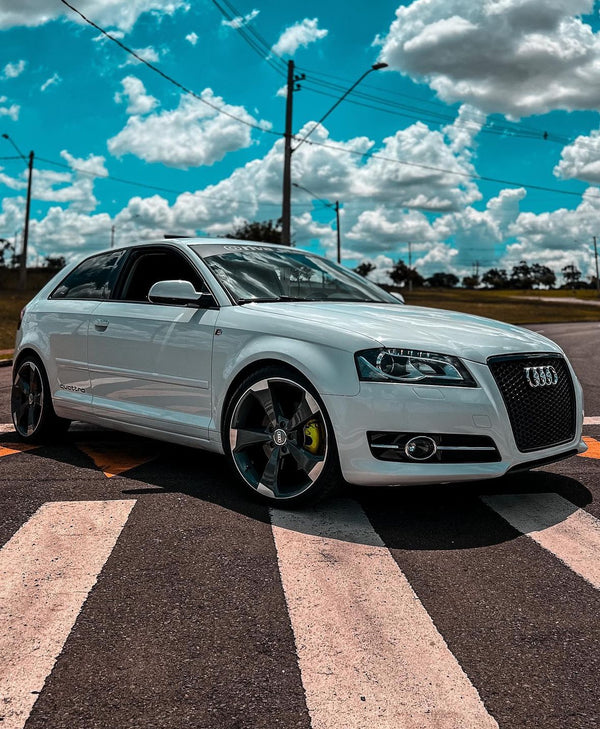 A white Audi car is parked diagonally on a road's crosswalk, under a partly cloudy blue sky. Power lines and grassy fields are visible in the background.