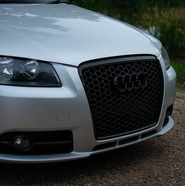 A silver car features a prominent black honeycomb grille with a logo of four rings, parked on a gravel path with greenery in the background.