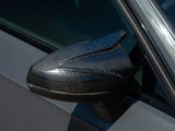 A carbon fiber car side mirror glistens in sunlight, attached to a grey vehicle door, reflecting its sleek, modern design.