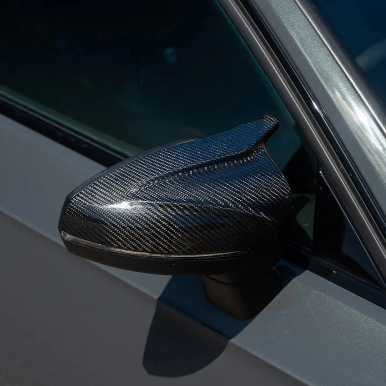 A carbon fiber car side mirror glistens in sunlight, attached to a grey vehicle door, reflecting its sleek, modern design.