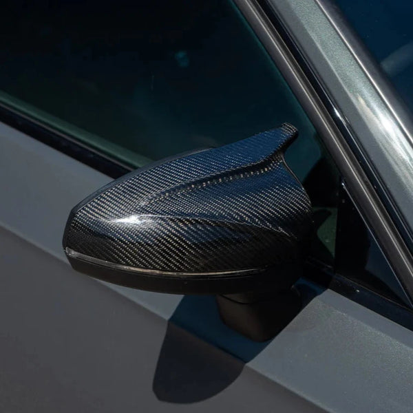 A carbon fiber car side mirror glistens in sunlight, attached to a grey vehicle door, reflecting its sleek, modern design.