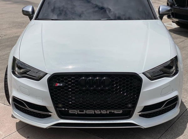A white Audi S3 car is parked on a paved area. The black front grille features the Audi logo and "quattro" text. The car's headlights are sleek and modern.