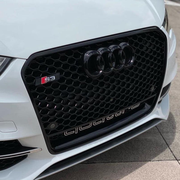Car grille with black honeycomb design features the Audi rings, an "S3" badge, and "quattro" text, set against the front of a white vehicle in an outdoor setting.