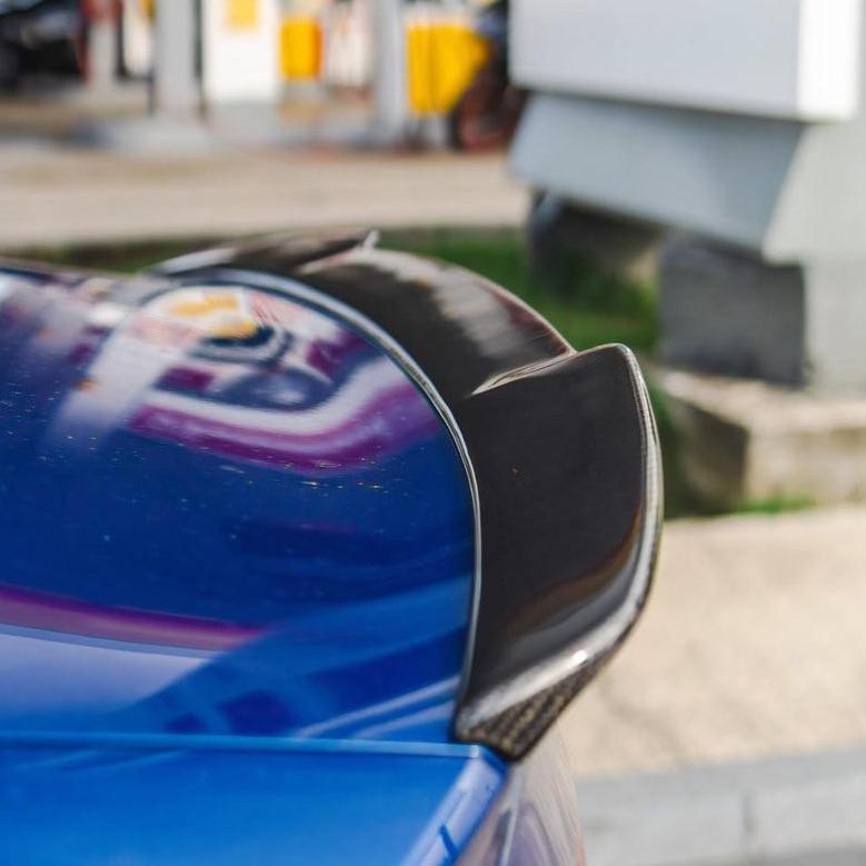A sleek, carbon fiber car spoiler is attached to a blue vehicle, parked near a building, with blurred urban elements in the background.
