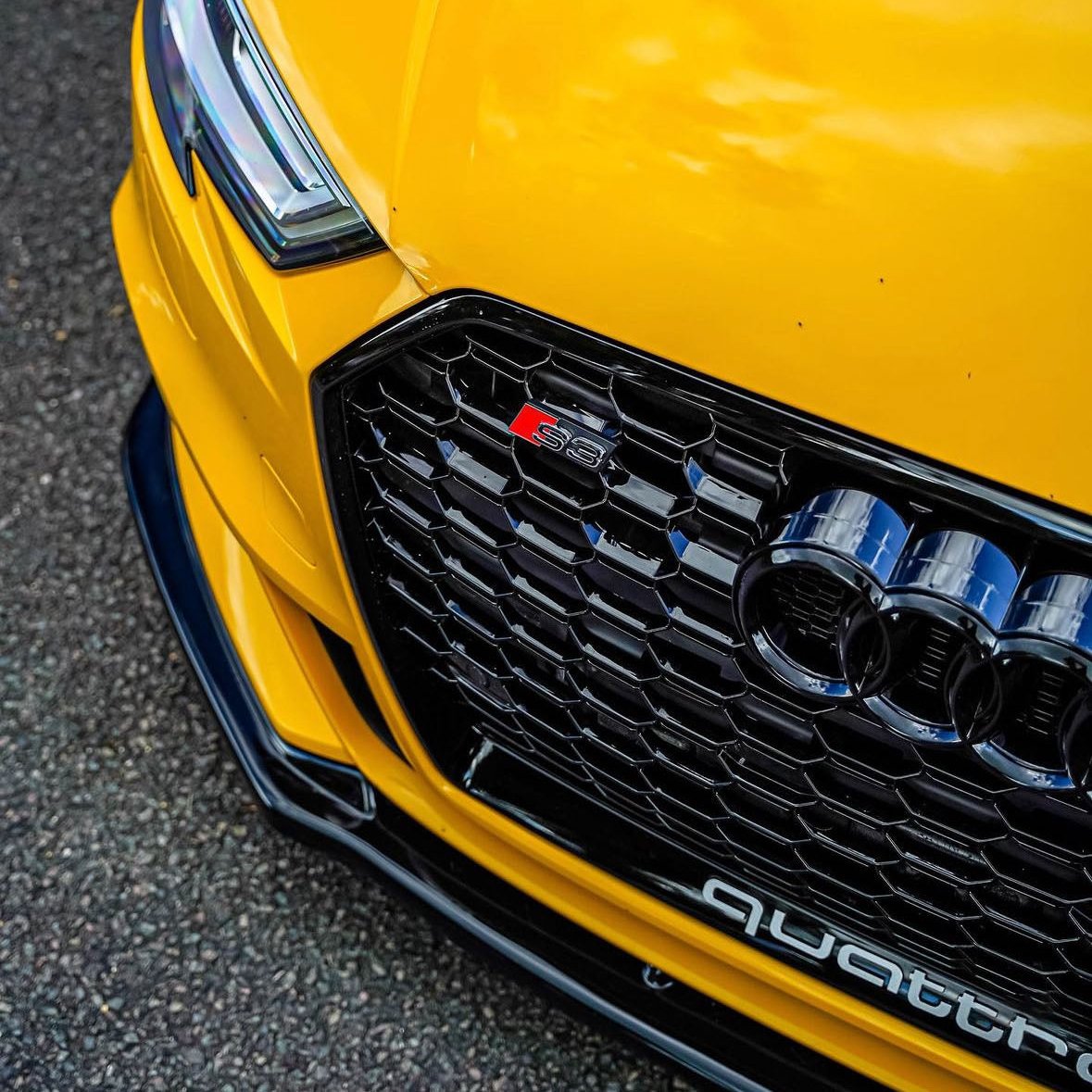 A yellow Audi S8 is parked on asphalt, featuring a prominent black grille with a red "S8" badge and the word "quattro" in white below.