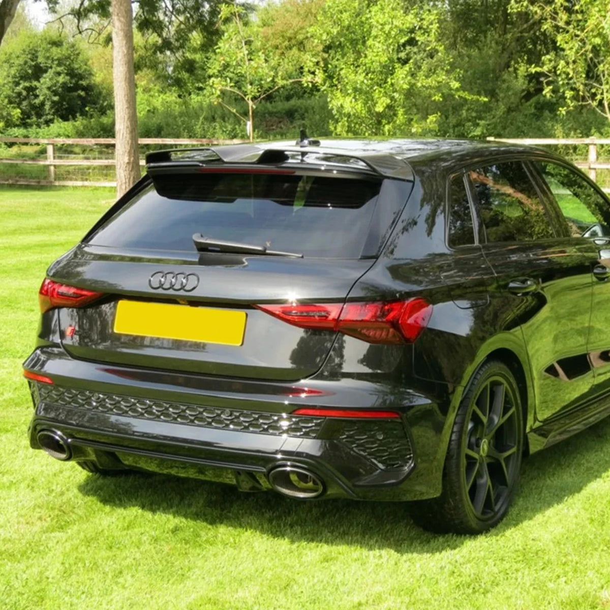 A black Audi car is parked on a grassy area surrounded by trees and a wooden fence. It has prominent dual exhaust pipes and tinted windows.
