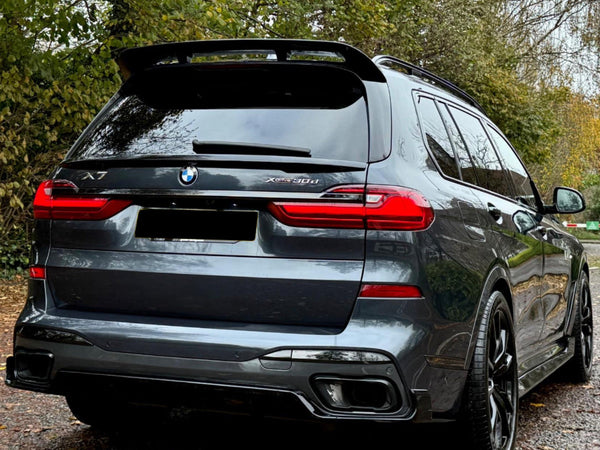 A dark gray BMW X7 parked on a leaf-covered path, surrounded by autumn trees. The car's rear features red taillights and dual exhaust. Visible text includes "X7" and "Xdrive30d."
