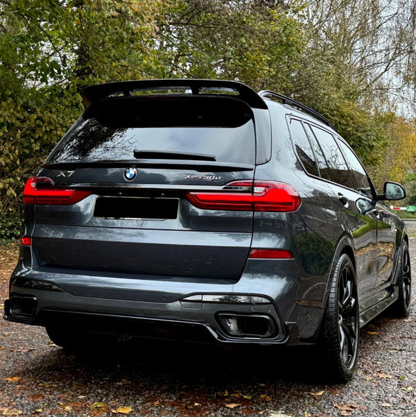A dark gray BMW X7 parked on a leaf-covered path, surrounded by autumn trees. The car's rear features red taillights and dual exhaust. Visible text includes "X7" and "Xdrive30d."