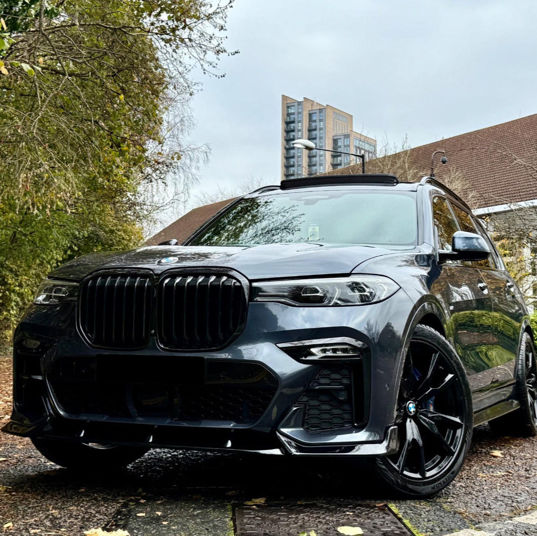 A sleek, black SUV is parked on a wet, leaf-covered road, surrounded by trees and buildings in the background, under a cloudy sky.
