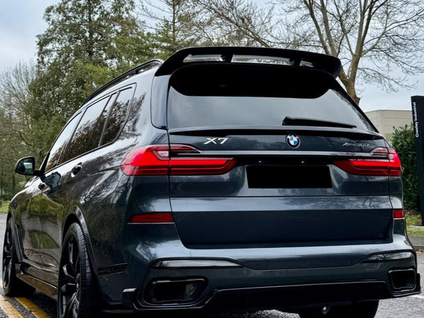 A dark-colored BMW X7 is parked on a road with double yellow lines. It features a prominent spoiler, surrounded by leafless trees and a cloudy sky.