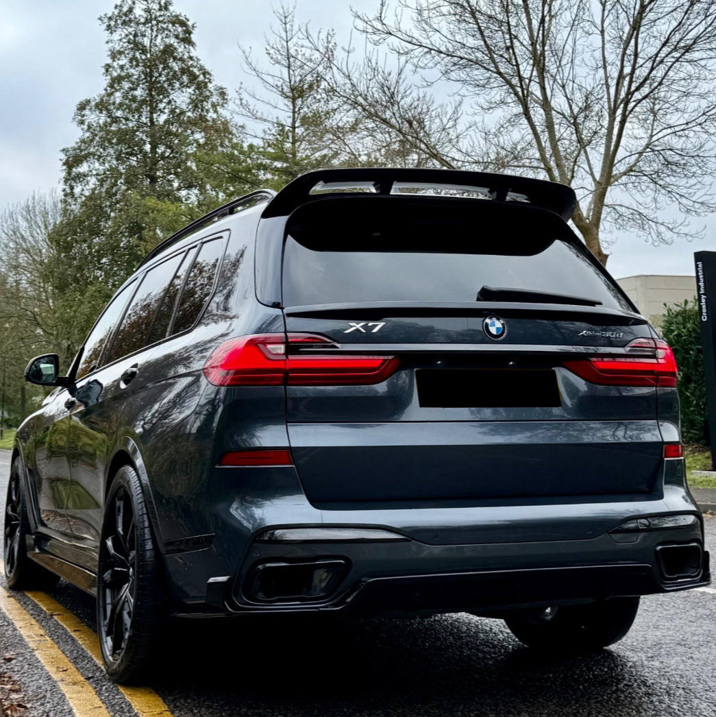 A dark-colored BMW X7 is parked on a road with double yellow lines. It features a prominent spoiler, surrounded by leafless trees and a cloudy sky.