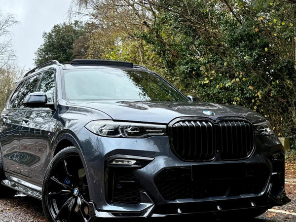 A gray SUV is parked on a gravel path, surrounded by lush green trees and overcast skies, showcasing its sleek front design and black accents.