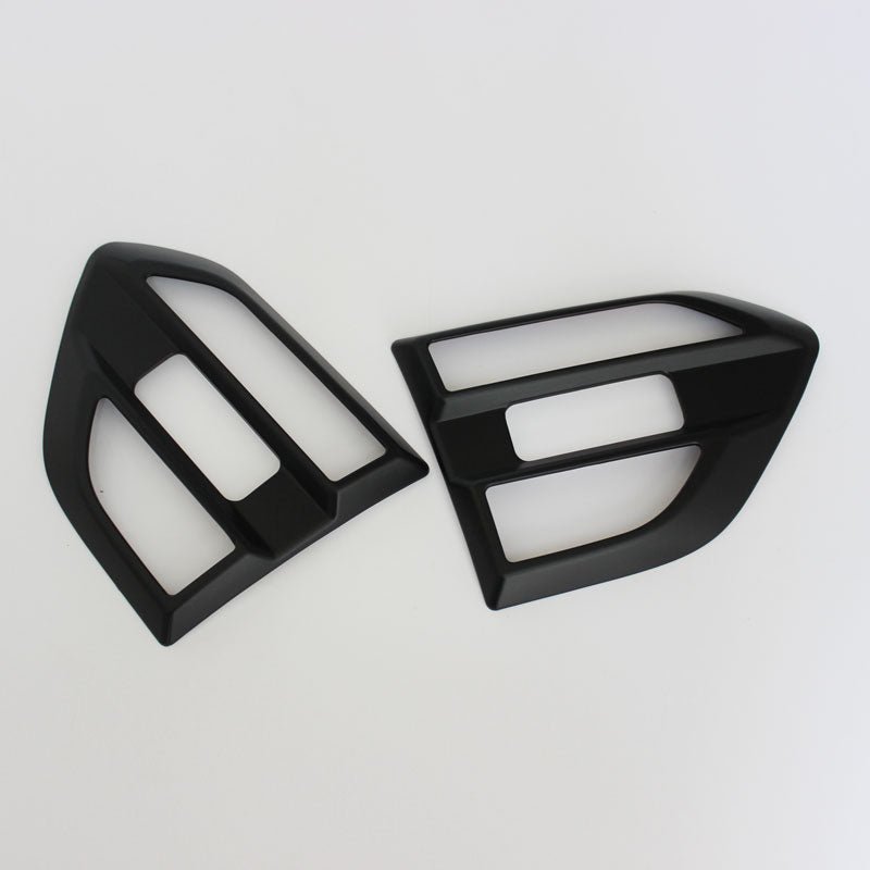 Two black plastic car components, resembling air vent covers, lie side by side on a white surface, showcasing a glossy finish and geometric cutouts.