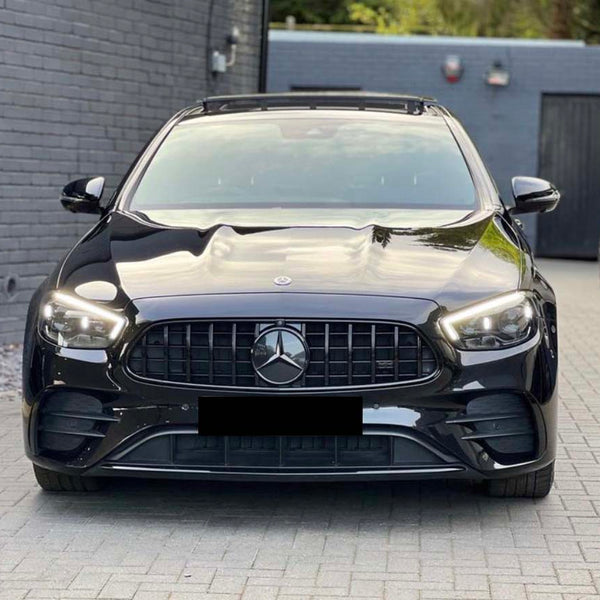 A black car with illuminated headlights is parked on a paved driveway. It's positioned in front of a gray brick wall, with another vehicle visible in the background.