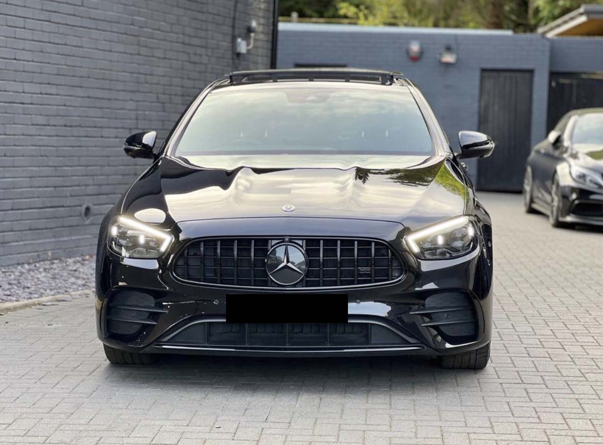 A black car with illuminated headlights is parked on a paved driveway. It's positioned in front of a gray brick wall, with another vehicle visible in the background.
