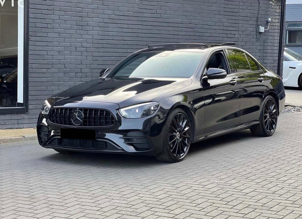 A black Mercedes sedan is parked on a gray brick driveway next to a modern building with dark walls and large windows. Another white car is partially visible in the background.