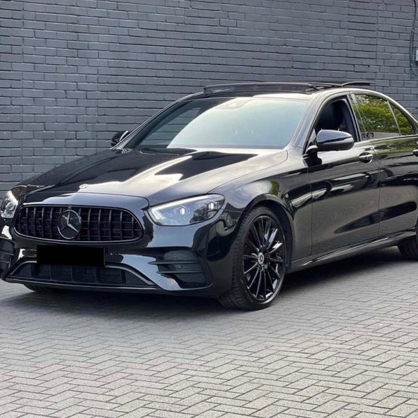 A black Mercedes sedan is parked on a gray brick driveway next to a modern building with dark walls and large windows. Another white car is partially visible in the background.