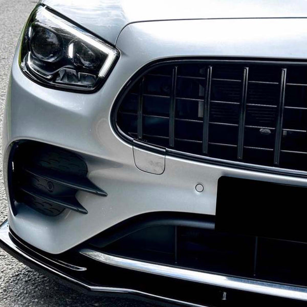 A silver car's front grille displays a prominent Mercedes-Benz emblem, parked on a gravel road with grass bordering the pavement.