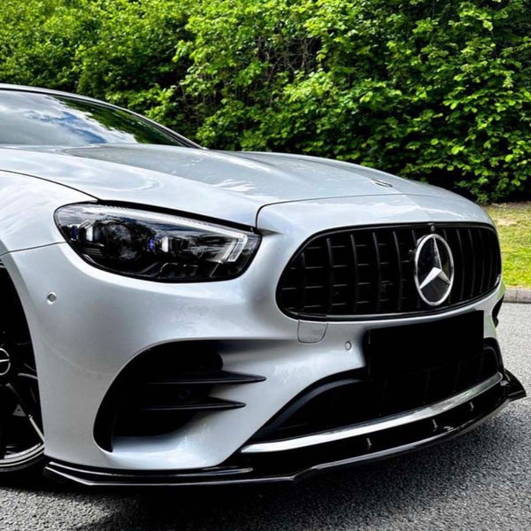 A silver car's front view with a sleek black grille and prominent emblem, parked on a gray road, set against a backdrop of dense green foliage.