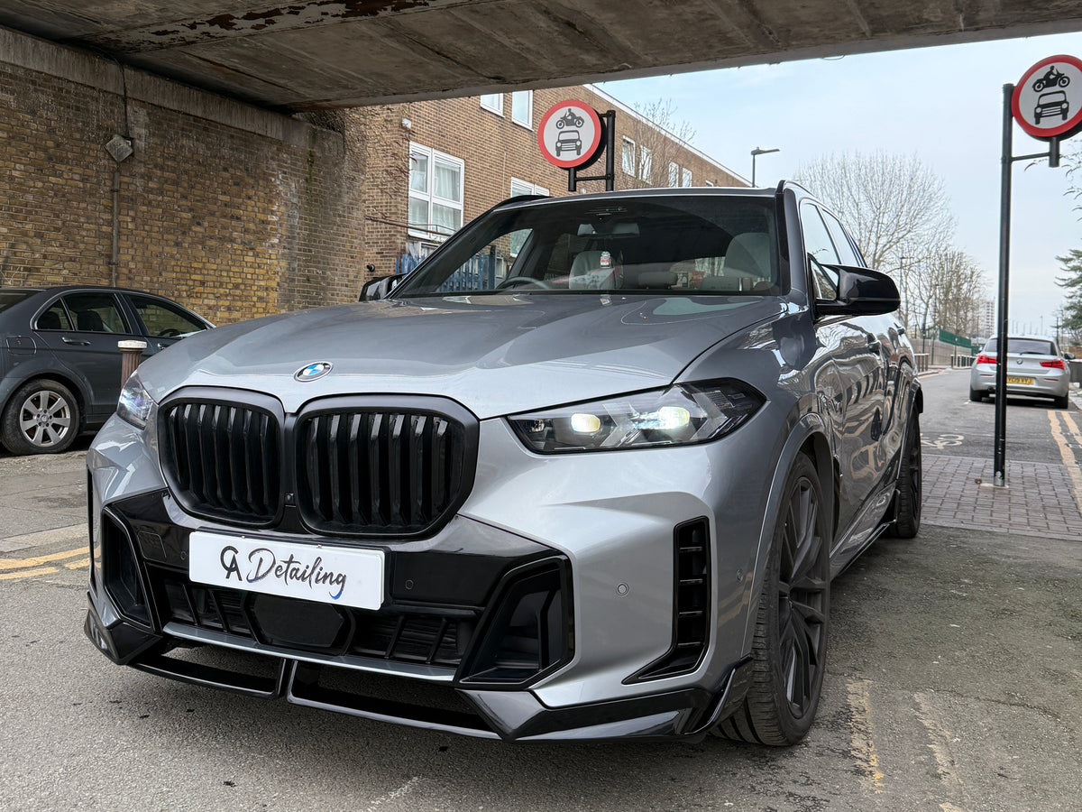 A silver BMW SUV is parked under a bridge with "A Detailing" on its front plate. Two "no cars allowed" signs are visible, surrounded by brick buildings and parked vehicles.