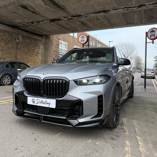 A silver BMW SUV is parked under a bridge with "A Detailing" on its front plate. Two "no cars allowed" signs are visible, surrounded by brick buildings and parked vehicles.